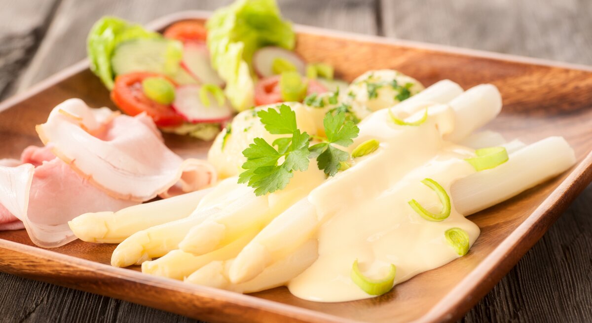 Seitliche Ansicht auf einen Teller mit Spargel und Schinken, begossen mit einer Sauce hollandaise. 