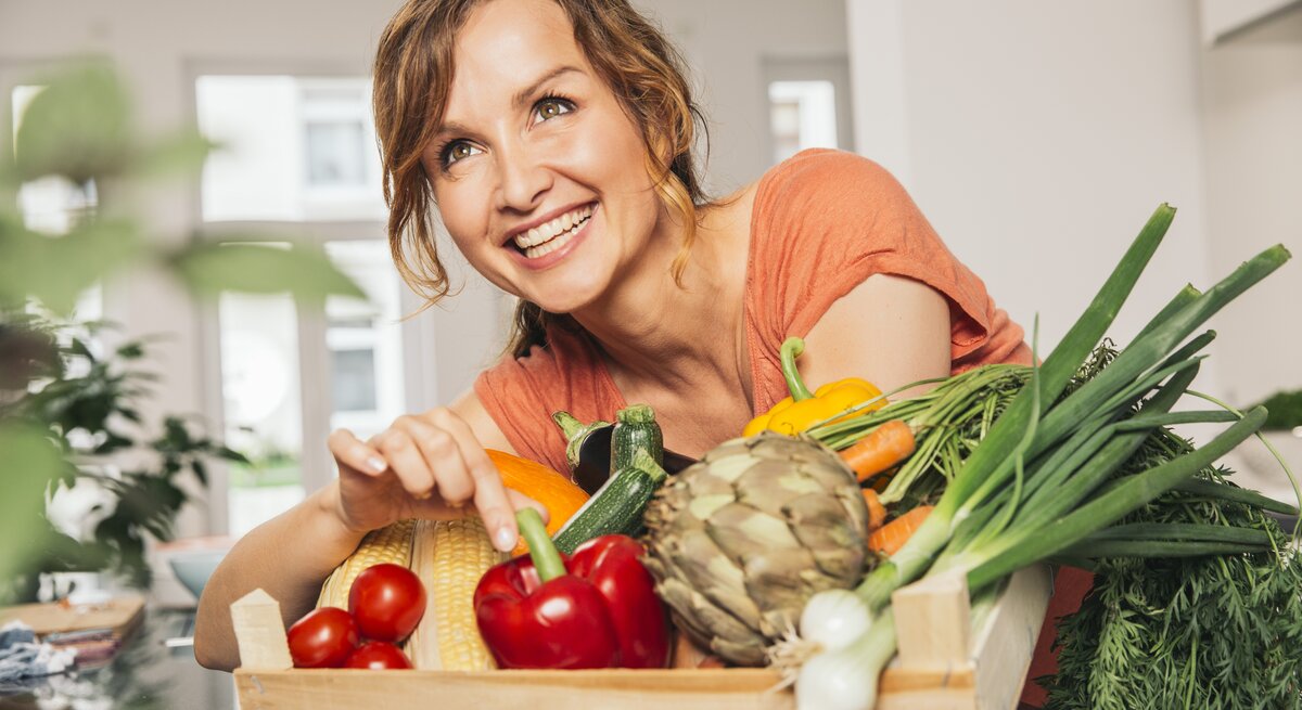 Eine strahlende Frau mit braunen, gelockten Haaren lehnt sich über eine Holzkiste voller frischer, bunter Gemüsesorten. Sie trägt ein korallenfarbenes Oberteil und zeigt mit einem Lächeln auf eine gelbe Paprika. Das Bild vermittelt eine fröhliche Stimmung und eine Leidenschaft für gesunde Ernährung. Sie befindet sich in einer modernen Küche, was auf eine häusliche und entspannte Atmosphäre hindeutet.