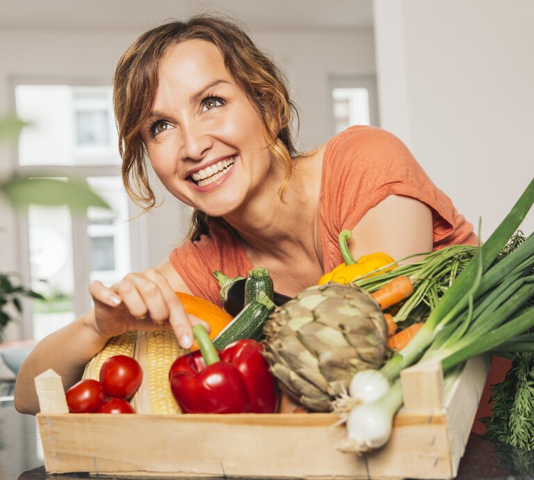 Eine strahlende Frau mit braunen, gelockten Haaren lehnt sich über eine Holzkiste voller frischer, bunter Gemüsesorten. Sie trägt ein korallenfarbenes Oberteil und zeigt mit einem Lächeln auf eine gelbe Paprika. Das Bild vermittelt eine fröhliche Stimmung und eine Leidenschaft für gesunde Ernährung. Sie befindet sich in einer modernen Küche, was auf eine häusliche und entspannte Atmosphäre hindeutet. | © Getty Images/Westend61