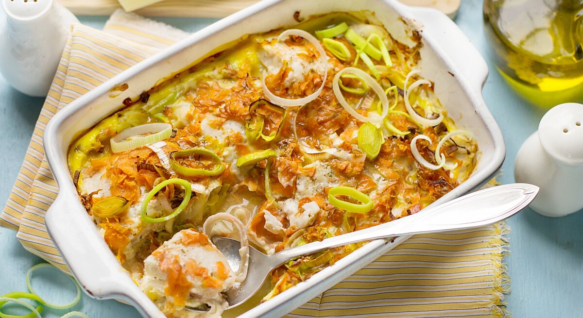 Ofengericht in weißer Auflaufform mit Fisch, Möhren und Lauch. Daneben Salzstreuer, eine Kanne Öl und ein Geschirrtuch.