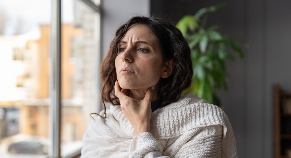 Eine kranke Frau steht zu Hause und berührt ihre geschwollenen Mandeln. Sie hat Schmerzen oder ein kratziges Gefühl im Hals. Die Frau leidet unter Angina oder Tonsillitis, verursacht durch Infektion oder Reizung.