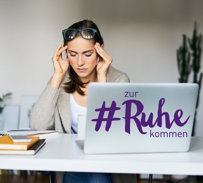 Eine junge Frau mit dunklen Haaren und einer Brille auf dem Kopf sitzt vor einem Laptop, auf dessen Deckel der Schriftzug "#zur Ruhe kommen" zu sehen ist. Sie hat die Augen geschlossen und ihre Finger an den Schläfen, was auf Stress oder Nachdenklichkeit hindeuten könnte. Vor ihr auf dem Tisch liegen ein Notizbuch, ein Tablet, ein Stapel bunter Haftnotizen und eine Tasse, was auf eine Arbeits- oder Lernsituation schließen lässt. Die Umgebung wirkt modern und hell. | © GettyImages/Westend61