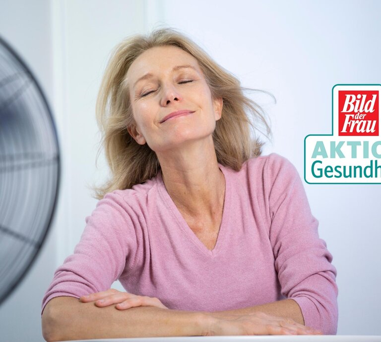 Eine reife Frau mit geschlossenen Augen und einem entspannten Lächeln genießt die kühle Brise eines Ventilators. Sie trägt ein rosa V-Ausschnitt-Shirt und lehnt sich in einem Stuhl zurück, ihre Arme locker auf dem Tisch vor ihr verschränkt. Ihr blondes Haar fällt locker um ihre Schultern. In der rechten oberen Ecke des Bildes ist ein Logo mit der Aufschrift "Bild der Frau AKTION Gesundheit" zu sehen. | © GettyImages/BSIPUIG [M]