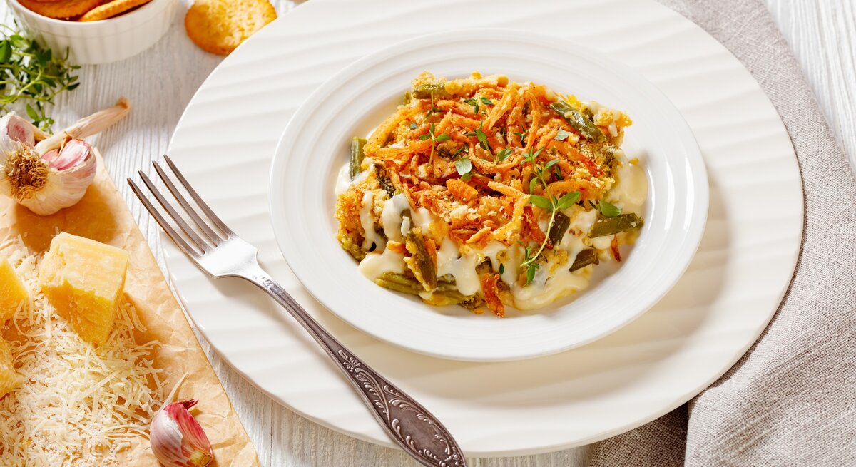 Bretonischer Bohnenauflauf auf weißem Teller mit Unterteller und Gabel auf hellem Untergrund. Im Bild Cracker, Parmesan und Knoblauch. Draufsicht.