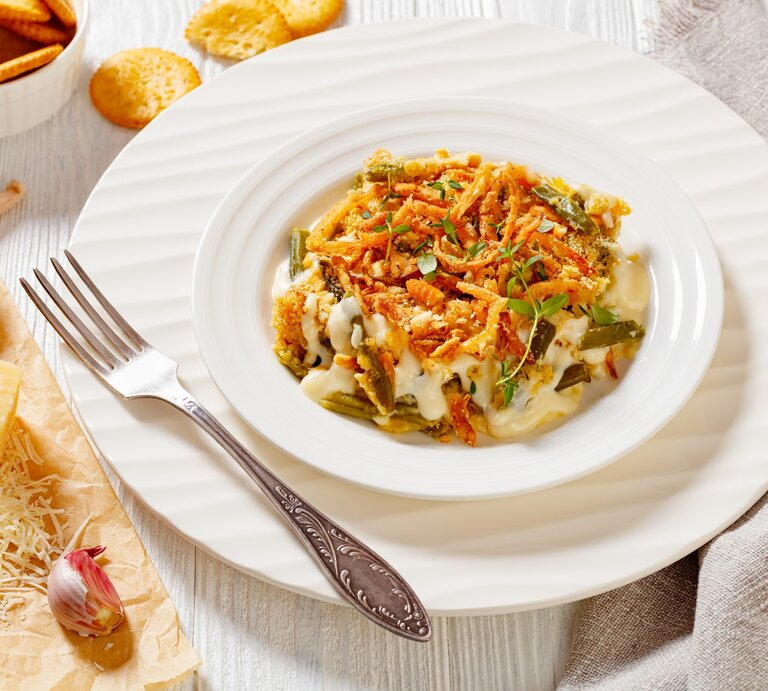 Bretonischer Bohnenauflauf auf weißem Teller mit Unterteller und Gabel auf hellem Untergrund. Im Bild Cracker, Parmesan und Knoblauch. Draufsicht. | © Getty Images / from_my_point_of_view