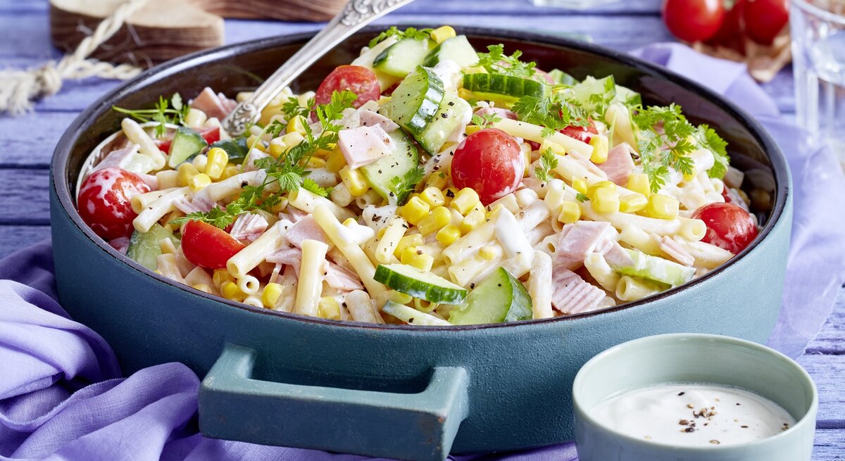 Nudelsalat mit Schinken in einer blauen Schüssel. Daneben ein Dressing und andere Zutaten. 