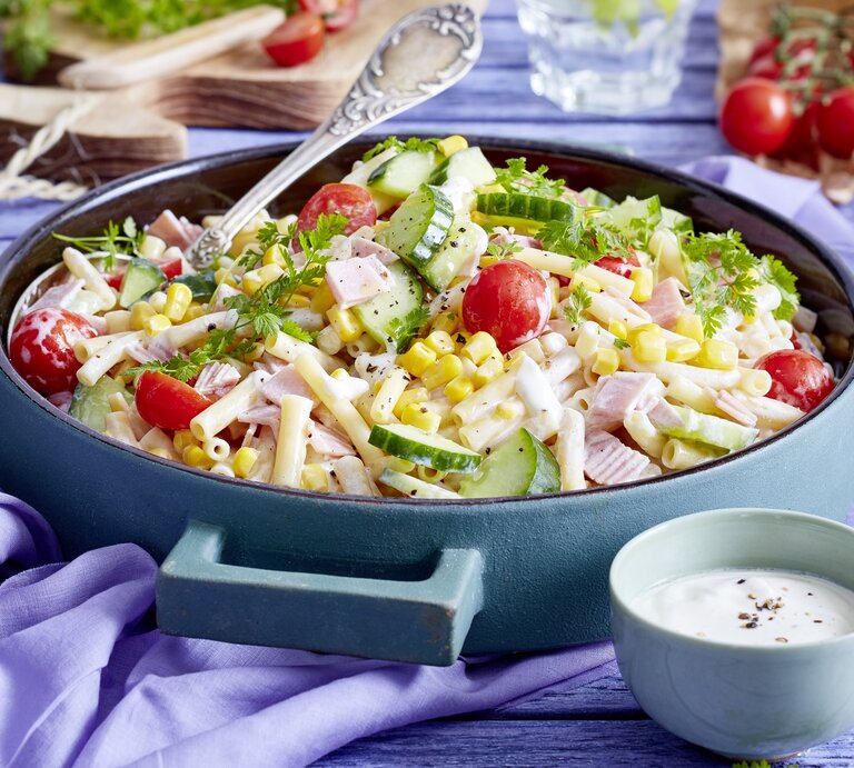 Nudelsalat mit Schinken in einer blauen Schüssel. Daneben ein Dressing und andere Zutaten.  | © EAT CLUB / Isabell Triemer 