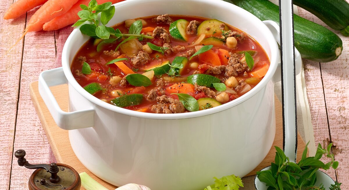Ein altmodischer weißer Topf gefüllt mit einer gesunden Suppe mit Gemüse und Hackfleisch. Um den Topf herum liegen Gemüsesorten. 
