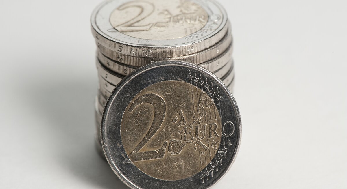 Das Bild zeigt eine Vorderansicht einer 2-Euro-Münze im Vordergrund, die auf ihrer Seite steht, mit einem Stapel weiterer 2-Euro-Münzen, die im Hintergrund unscharf zu sehen sind. Die Münze im Vordergrund hat einen silberfarbenen Kern und einen goldfarbenen Rand. Auf ihr ist das Nominal "2 EURO" zu erkennen, umgeben von einer geographischen Darstellung Europas und Sternen, die die Europäische Union symbolisieren.
