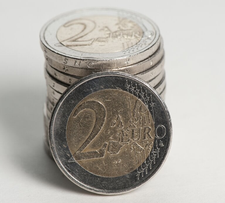 Das Bild zeigt eine Vorderansicht einer 2-Euro-Münze im Vordergrund, die auf ihrer Seite steht, mit einem Stapel weiterer 2-Euro-Münzen, die im Hintergrund unscharf zu sehen sind. Die Münze im Vordergrund hat einen silberfarbenen Kern und einen goldfarbenen Rand. Auf ihr ist das Nominal "2 EURO" zu erkennen, umgeben von einer geographischen Darstellung Europas und Sternen, die die Europäische Union symbolisieren. | © iStock.com / Photomick