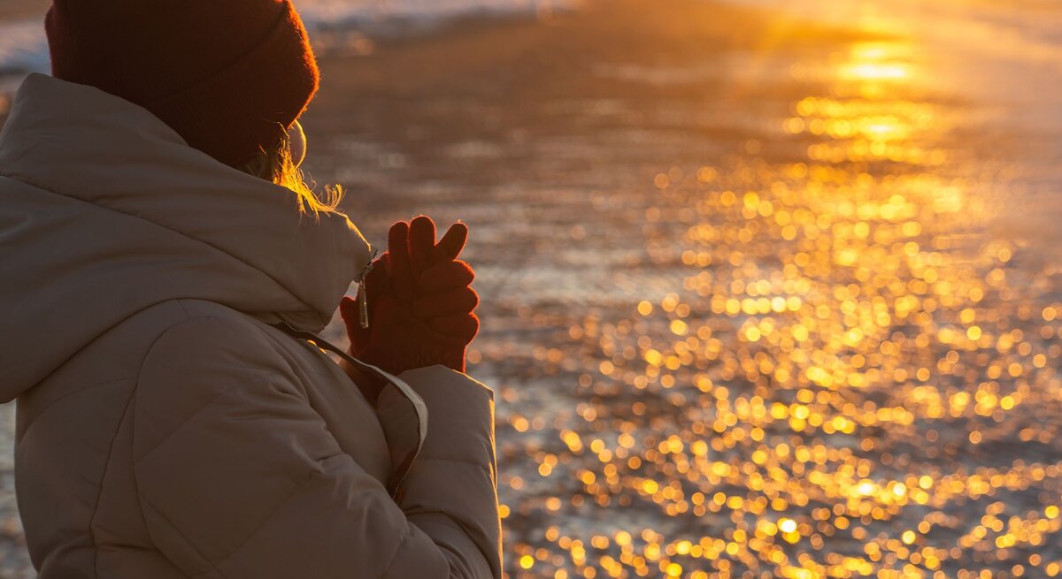 Eine Frau mit beigem Wintermantel und roter Mütze steht bei Sonnenuntergang am Strand und wärmt ihre Hände.
