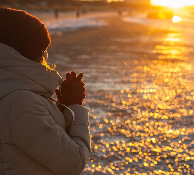 Eine Frau mit beigem Wintermantel und roter Mütze steht bei Sonnenuntergang am Strand und wärmt ihre Hände. | ©  iStock/SrdjanPav
