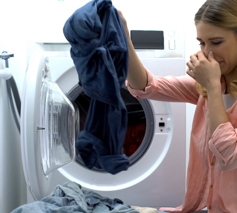 Frau holt mit der einen Hand Wäsche aus der Waschmaschine und hält sich mit der anderen die Nase zu | © iStock.com / Motortion