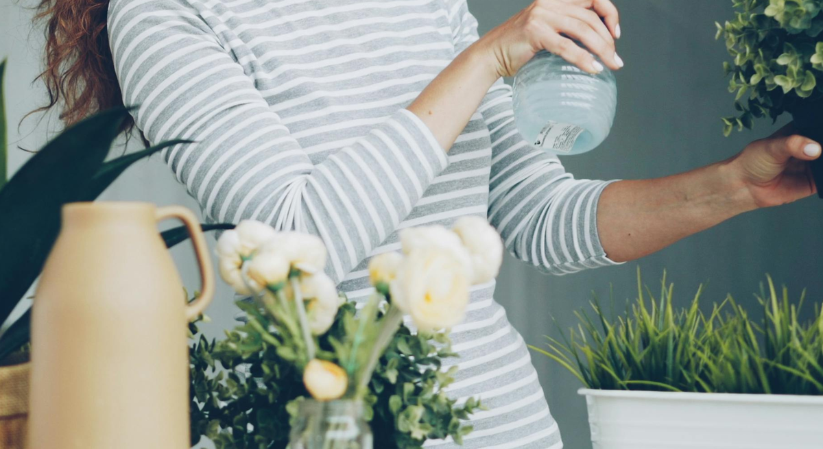 Eine Frau mit lockigem rötlichem Haar sprüht in Freizeitkleidung Wasser auf eine grüne Pflanze, umgeben von Vasen und Blumen.