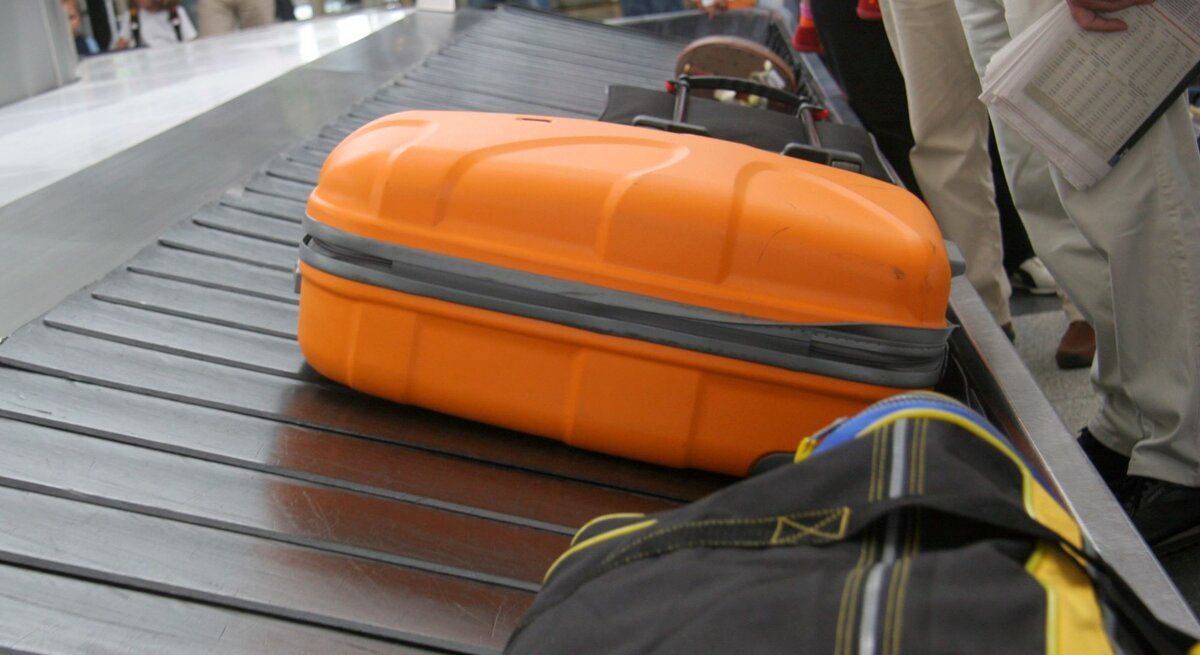  Das Bild zeigt einen Abschnitt eines Gepäckausgabebands an einem Flughafen aus einer niedrigen Perspektive. Im Vordergrund ist ein auffälliger orangefarbener Koffer zu sehen, der sich auf dem Band bewegt. Im Hintergrund sind unscharf weitere Gepäckstücke und wartende Passagiere zu erkennen. Die Menschen stehen oder gehen entlang des Bands, wahrscheinlich in Erwartung ihres eigenen Gepäcks. Der Fokus liegt auf dem orangefarbenen Koffer, was darauf hindeutet, dass die Aufnahme aus der Sicht eines wartenden Passagiers gemacht wurde.