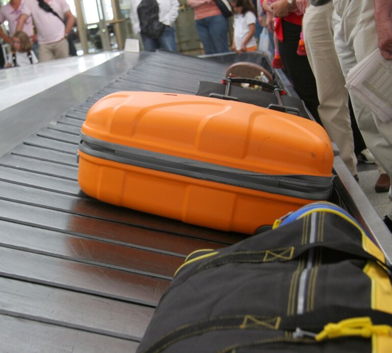  Das Bild zeigt einen Abschnitt eines Gepäckausgabebands an einem Flughafen aus einer niedrigen Perspektive. Im Vordergrund ist ein auffälliger orangefarbener Koffer zu sehen, der sich auf dem Band bewegt. Im Hintergrund sind unscharf weitere Gepäckstücke und wartende Passagiere zu erkennen. Die Menschen stehen oder gehen entlang des Bands, wahrscheinlich in Erwartung ihres eigenen Gepäcks. Der Fokus liegt auf dem orangefarbenen Koffer, was darauf hindeutet, dass die Aufnahme aus der Sicht eines wartenden Passagiers gemacht wurde. | © Adobe Stock/ axeldrosta