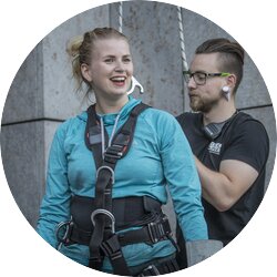 Eine lächelnde Frau in Kletterausrüstung und blauer Sportkleidung steht neben einem Mann mit Brille und Kopfhörern. Sie scheint sich auf eine Kletteraktivität vorzubereiten, während der Mann im Hintergrund steht und ein Sicherungsgerät hält, beide vor einer grauen Betonwand | © Pascal Rohe