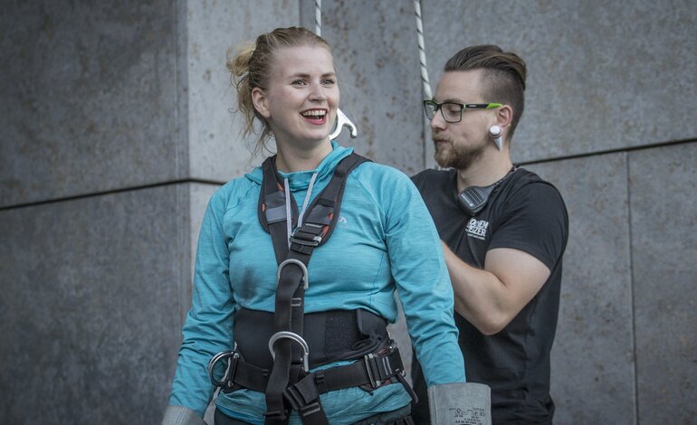 Eine lächelnde Frau in Kletterausrüstung und blauer Sportkleidung steht neben einem Mann mit Brille und Kopfhörern. Sie scheint sich auf eine Kletteraktivität vorzubereiten, während der Mann im Hintergrund steht und ein Sicherungsgerät hält, beide vor einer grauen Betonwand | © Pascal Rohe