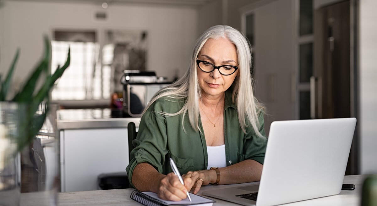 Eine konzentrierte ältere Frau mit langen grauen Haaren und Brille sitzt an einem hellen Arbeitsplatz und schreibt etwas in ein Notizbuch. Vor ihr steht ein geöffneter Laptop. Sie trägt ein olivgrünes Hemd und wirkt, als wäre sie gerade in einer Arbeits- oder Lernumgebung zu Hause. Das Zimmer ist hell und modern eingerichtet, mit einer Küche im Hintergrund, was auf ein häusliches Büro hindeutet. Das natürliche Licht und die Pflanze auf dem Tisch schaffen eine angenehme und ruhige Arbeitsatmosphäre.