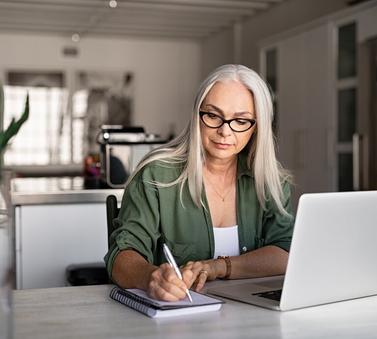 Eine konzentrierte ältere Frau mit langen grauen Haaren und Brille sitzt an einem hellen Arbeitsplatz und schreibt etwas in ein Notizbuch. Vor ihr steht ein geöffneter Laptop. Sie trägt ein olivgrünes Hemd und wirkt, als wäre sie gerade in einer Arbeits- oder Lernumgebung zu Hause. Das Zimmer ist hell und modern eingerichtet, mit einer Küche im Hintergrund, was auf ein häusliches Büro hindeutet. Das natürliche Licht und die Pflanze auf dem Tisch schaffen eine angenehme und ruhige Arbeitsatmosphäre. | © iStock.com/Ridofranz