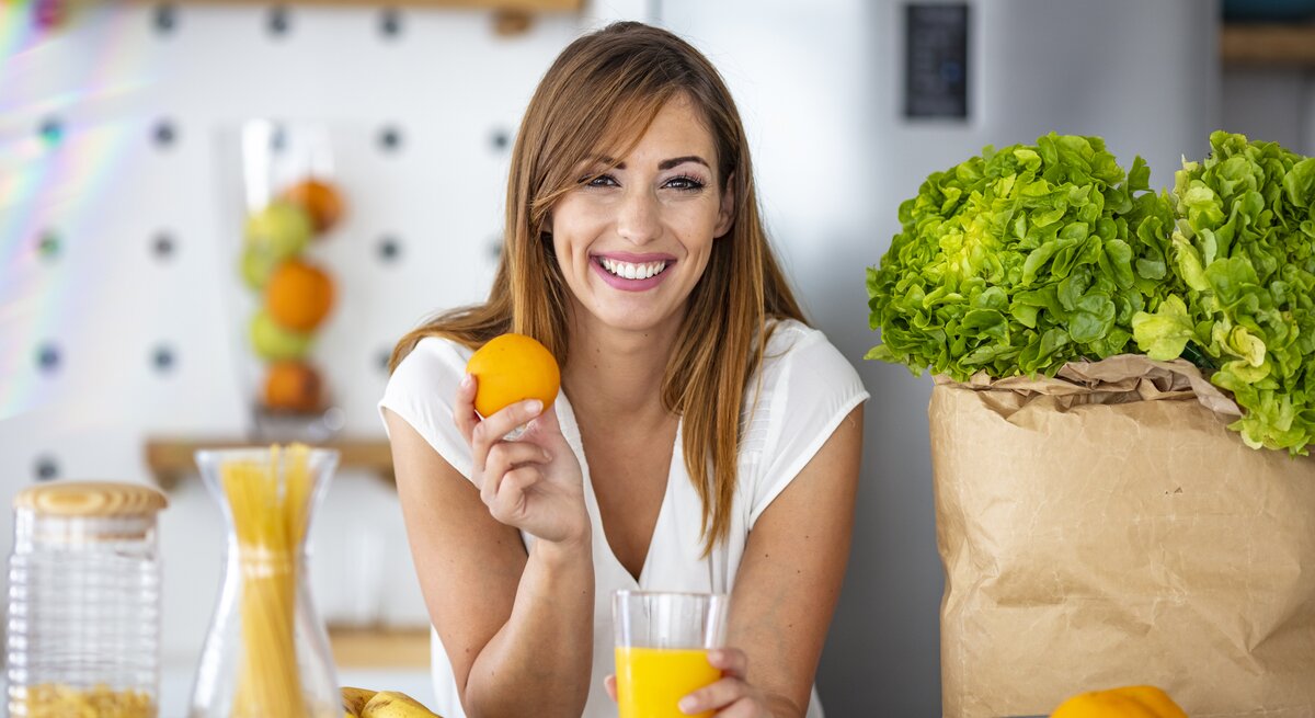 Das Bild zeigt eine fröhliche, junge Frau in einer modernen Küche, die eine Orange in der Hand hält und ein Glas Orangensaft auf dem Tisch vor sich hat. Sie hat langes, brünettes Haar und trägt ein weißes T-Shirt. Im Hintergrund sind eine Vase mit Spaghetti, eine Tüte mit frischem Salatkopf und eine Schüssel mit Zitrusfrüchten zu sehen. Die Frau lächelt in die Kamera, was die Assoziation von Gesundheit und Freude mit dem Verzehr von frischen Früchten und einer ausgewogenen Ernährung vermittelt.