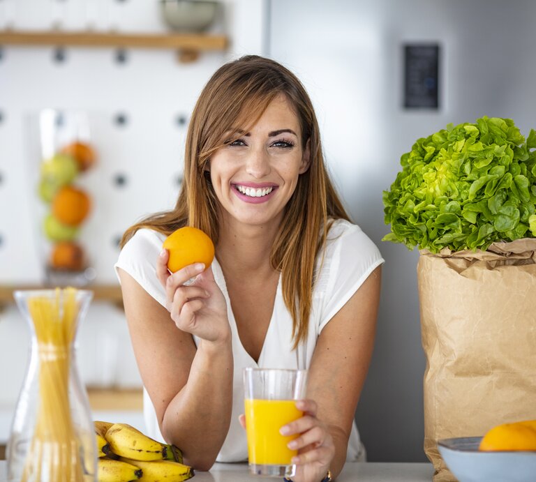 Das Bild zeigt eine fröhliche, junge Frau in einer modernen Küche, die eine Orange in der Hand hält und ein Glas Orangensaft auf dem Tisch vor sich hat. Sie hat langes, brünettes Haar und trägt ein weißes T-Shirt. Im Hintergrund sind eine Vase mit Spaghetti, eine Tüte mit frischem Salatkopf und eine Schüssel mit Zitrusfrüchten zu sehen. Die Frau lächelt in die Kamera, was die Assoziation von Gesundheit und Freude mit dem Verzehr von frischen Früchten und einer ausgewogenen Ernährung vermittelt. | © iStockcom/dragana991