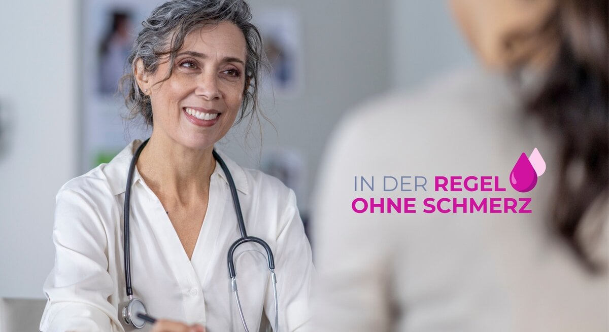 Eine freundlich lächelnde Ärztin mittleren Alters mit grauen Locken und einem Stethoskop um den Hals steht in einem hellen medizinischen Untersuchungsraum. Sie trägt einen weißen Arztkittel und scheint eine Patientin zu begrüßen, die außerhalb des Bildes sitzt und die die Ärztin anschaut. Im Vordergrund ist der Schriftzug "IN DER REGEL OHNE SCHMERZ" in lila Farbe mit einem rosa Tropfensymbol zu sehen.