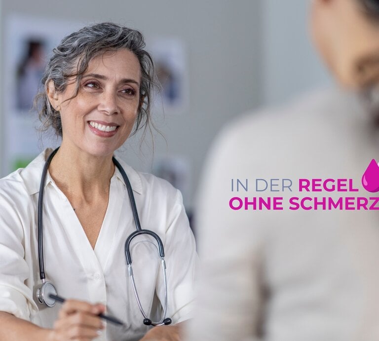 Eine freundlich lächelnde Ärztin mittleren Alters mit grauen Locken und einem Stethoskop um den Hals steht in einem hellen medizinischen Untersuchungsraum. Sie trägt einen weißen Arztkittel und scheint eine Patientin zu begrüßen, die außerhalb des Bildes sitzt und die die Ärztin anschaut. Im Vordergrund ist der Schriftzug "IN DER REGEL OHNE SCHMERZ" in lila Farbe mit einem rosa Tropfensymbol zu sehen. | © GettyImages/FatCamera
