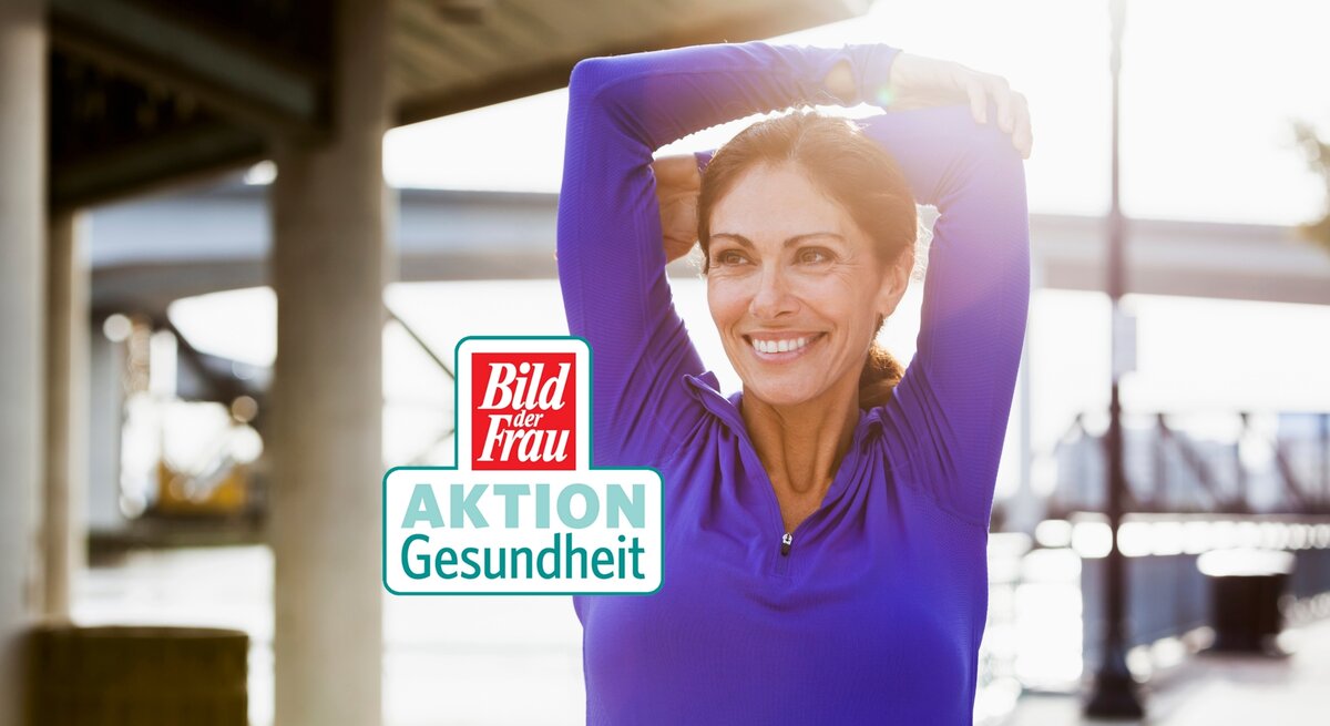 Eine lächelnde Frau mittleren Alters in einem leuchtend blauen Sportshirt dehnt sich draußen, indem sie ihre Arme hinter dem Kopf verschränkt. Sie strahlt Gesundheit und Zufriedenheit aus und genießt sichtlich ihre Fitnessroutine. Im Hintergrund ist eine unscharfe städtische Umgebung zu erkennen. Im Vordergrund ist ein Logo mit den Worten "Bild der Frau AKTION Gesundheit" zu sehen.
