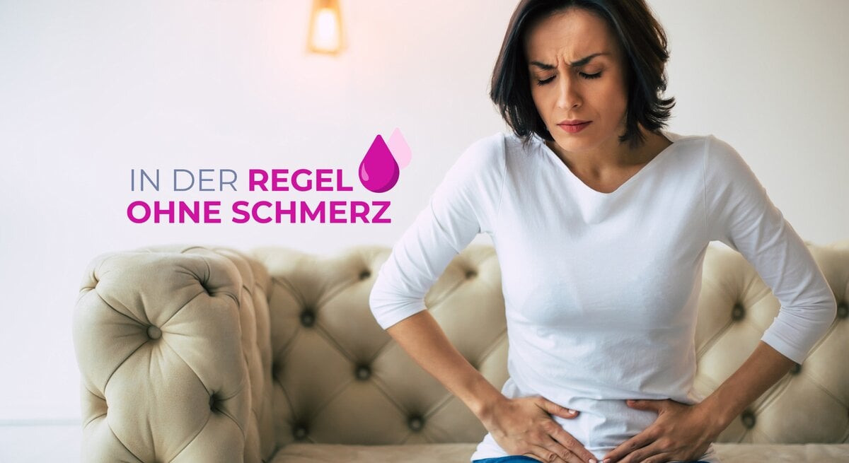 Eine Frau mittleren Alters mit dunklen Haaren sitzt auf einer cremefarbenen Couch und hält sich den Unterbauch, während sie schmerzverzerrt nach unten blickt. Sie trägt ein schlichtes weißes Oberteil und blaue Jeans. Ihr Gesichtsausdruck deutet auf Unbehagen und Bauchschmerzen hin. Über ihr schwebt der Text "IN DER REGEL OHNE SCHMERZ" in Großbuchstaben, begleitet von einem Tropfensymbol. Der Hintergrund ist hell und modern gestaltet, mit dezenten Wandleuchten, die eine warme und einladende Atmosphäre erzeugen.