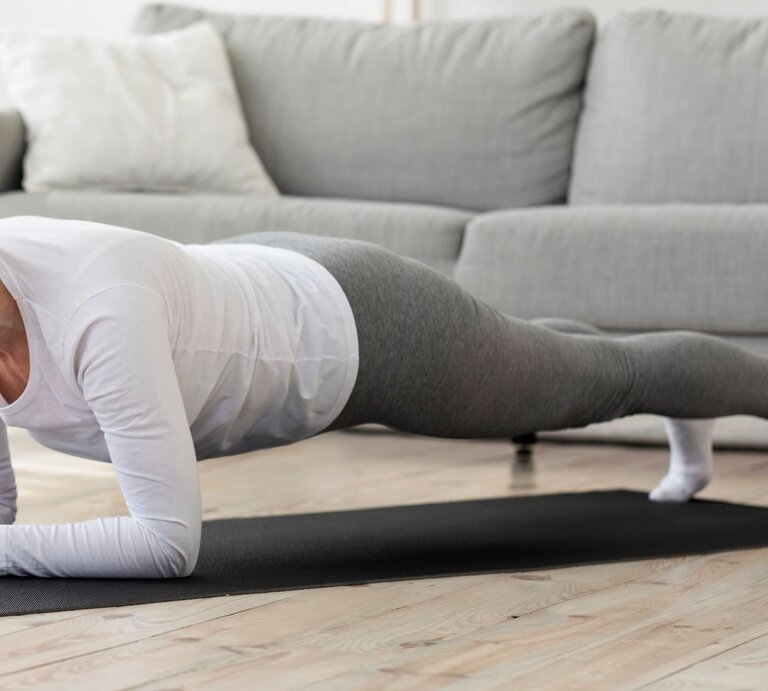 Ältere Frau, die eine Plank-Übung macht. Sie trägt ein weißes Langarmshirt, graue Leggings und weiße Socken. Die Person befindet sich in einer horizontalen Position mit dem Körper gerade und gestreckt, gestützt auf die Unterarme und Zehen. Im Hintergrund ein Wohnzimmer mit Sofa und neutralem Farbschema. | © Getty Images / Prostock-Studio