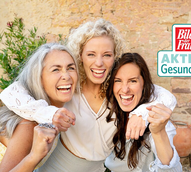 Drei lachende Frauen unterschiedlichen Alters, die Freude und Zusammenhalt ausstrahlen, stehen eng beieinander und blicken direkt in die Kamera. Die ältere Frau mit grauen Haaren und die zwei jüngeren Frauen mit blonden und braunen Haaren zeigen mit ihren Fäusten symbolisch ihre Stärke und Energie. Sie tragen leichte, lässige Kleidung und befinden sich vor einer natürlichen Steinfassade mit grünen Pflanzen im Hintergrund. In der oberen rechten Ecke des Bildes ist das Logo "Bild der Frau AKTION Gesundheit" zu sehen, was das Engagement für weibliche Gesundheit und Wohlbefinden darstellt. | © GettyImages/Westend61