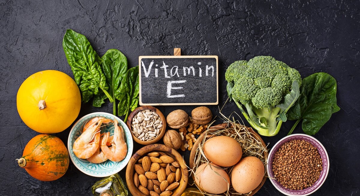 Das Bild zeigt eine Zusammenstellung von Lebensmitteln, die reich an Vitamin E sind, angeordnet auf einer dunklen Oberfläche. Im Vordergrund befindet sich eine kleine Schiefertafel mit der Aufschrift "Vitamin E". Zu den dargestellten Nahrungsmitteln gehören Blattgemüse, Nüsse, Samen, ein Kürbis, Brokkoli, Shrimps und Eier. Das Bild betont die Bedeutung einer ausgewogenen Ernährung mit Fokus auf Nährstoffen, die für die Gesundheit der Haut und allgemeines Wohlbefinden wichtig sind.