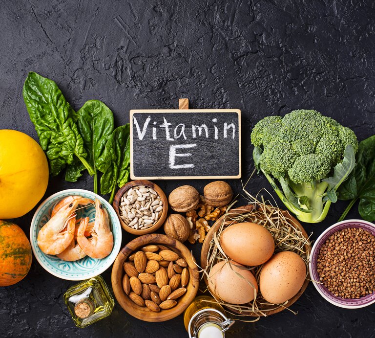 Das Bild zeigt eine Zusammenstellung von Lebensmitteln, die reich an Vitamin E sind, angeordnet auf einer dunklen Oberfläche. Im Vordergrund befindet sich eine kleine Schiefertafel mit der Aufschrift "Vitamin E". Zu den dargestellten Nahrungsmitteln gehören Blattgemüse, Nüsse, Samen, ein Kürbis, Brokkoli, Shrimps und Eier. Das Bild betont die Bedeutung einer ausgewogenen Ernährung mit Fokus auf Nährstoffen, die für die Gesundheit der Haut und allgemeines Wohlbefinden wichtig sind. | © GettyImages/yulka3ice