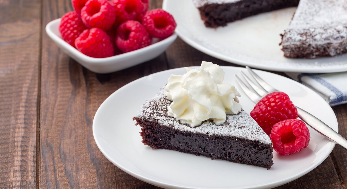 Schokoladenkuchen mit Puderzucker, Sahne und Himbeeren auf weißem Teller in rustikalem Ambiente. 