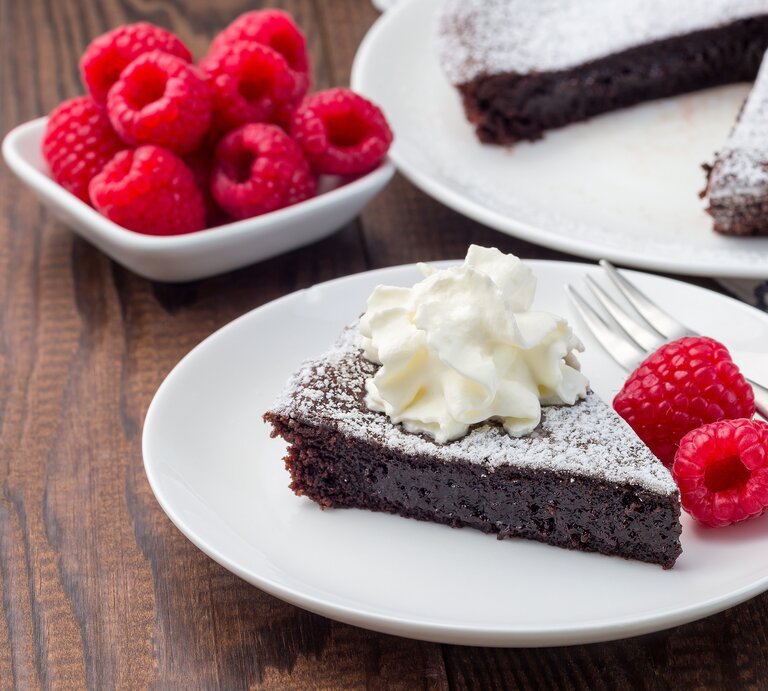 Schokoladenkuchen mit Puderzucker, Sahne und Himbeeren auf weißem Teller in rustikalem Ambiente.  | © Getty Images/iuliia_n