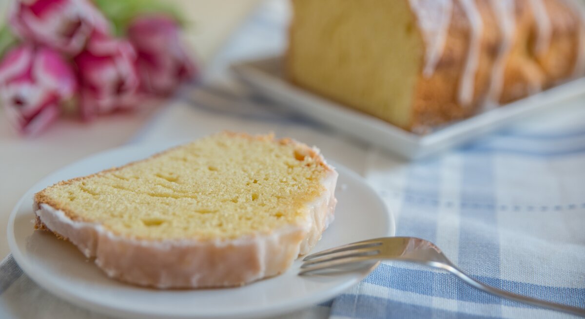 Ein Stück vom Zitronenkuchen auf einem weißen Teller mit hellblau-weiß-karierter Tischdecke. Alles nah und von der Seite fotografiert.
