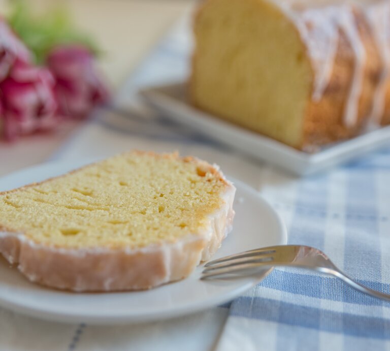 Ein Stück vom Zitronenkuchen auf einem weißen Teller mit hellblau-weiß-karierter Tischdecke. Alles nah und von der Seite fotografiert. | © iStock/ A_Lein