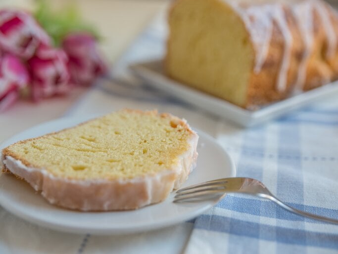Ein Stück vom Zitronenkuchen auf einem weißen Teller mit hellblau-weiß-karierter Tischdecke. Alles nah und von der Seite fotografiert. | © iStock/ A_Lein