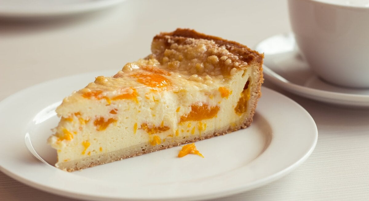 Ein angeschnittenes Stück Mandarinen-Schmandkuchen auf weißem Teller. 