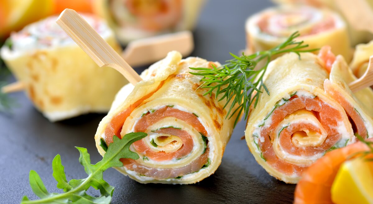 Dunkler Untergrund. Darauf liegen aufgerollte Pfannkuchen mit einer Füllung aus Lachs und Frischkäse.