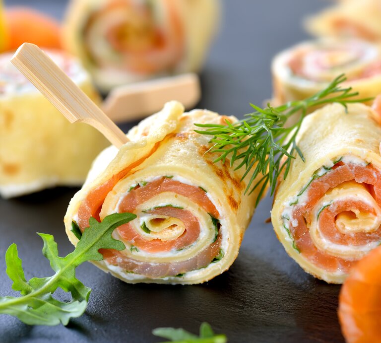 Dunkler Untergrund. Darauf liegen aufgerollte Pfannkuchen mit einer Füllung aus Lachs und Frischkäse. | © iStock/ kabVisio