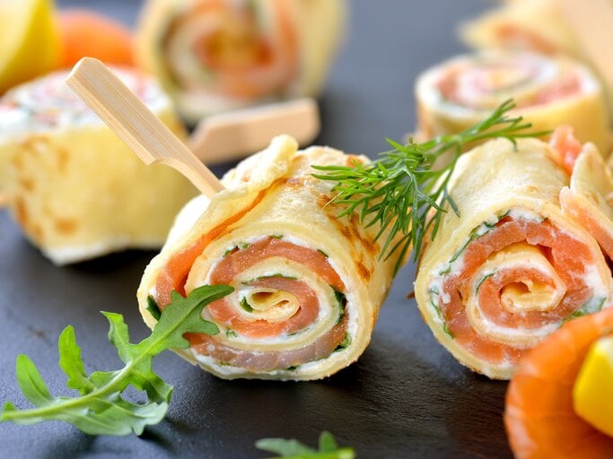 Dunkler Untergrund. Darauf liegen aufgerollte Pfannkuchen mit einer Füllung aus Lachs und Frischkäse. | © iStock/ kabVisio