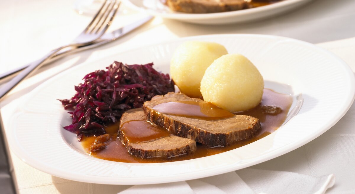 Rheinischer Sauerbraten mit Rotkohl und Kartoffelklößen auf weißem Teller. 