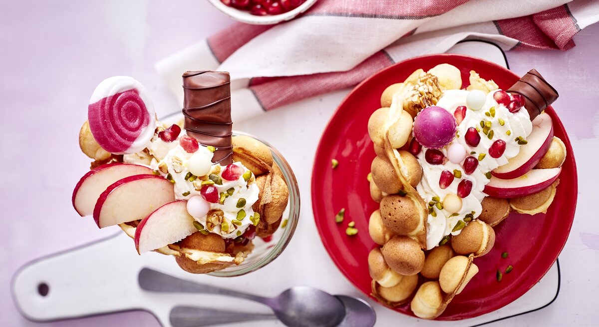 Eine große Bubble-Waffel auf einem runden Teller / eine kleine im Glas / zwei Teelöffel / Brettchen / Küchentuch / Draufsicht