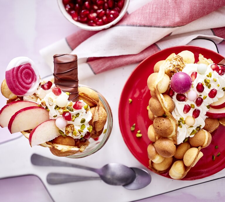 Eine große Bubble-Waffel auf einem runden Teller / eine kleine im Glas / zwei Teelöffel / Brettchen / Küchentuch / Draufsicht | © Eat Club; Isabell Triemer