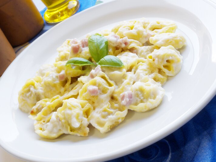 Auf dem Bild ist ein Teller mit Tortellini in einer cremigen Sauce zu sehen. Die Pasta ist mit kleinen Schinkenwürfeln angereichert und mit frischem Basilikum garniert. Der Teller steht auf einem Tisch mit einer blauen Tischdecke und im Hintergrund ist eine Flasche Olivenöl angedeutet. Das Gericht wirkt frisch und ist klassisch italienisch angerichtet. | © iStock/ NiroDesign