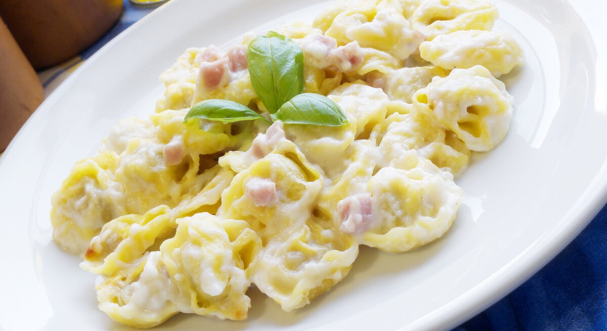 Auf dem Bild ist ein Teller mit Tortellini in einer cremigen Sauce zu sehen. Die Pasta ist mit kleinen Schinkenwürfeln angereichert und mit frischem Basilikum garniert. Der Teller steht auf einem Tisch mit einer blauen Tischdecke und im Hintergrund ist eine Flasche Olivenöl angedeutet. Das Gericht wirkt frisch und ist klassisch italienisch angerichtet.
