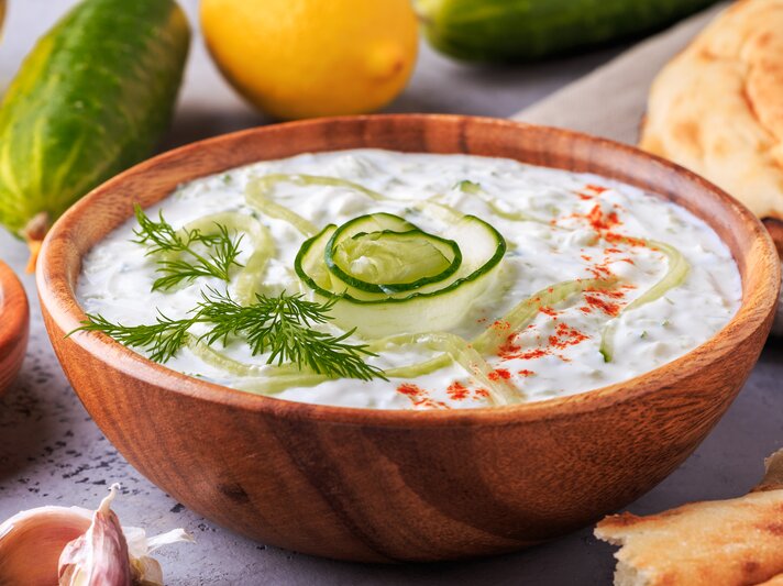 Frischer Zaziki mit Gurke in einer Holzschale. Daneben liegen Gurken, Knoblauch, Zitronen und Brot.  | © iStock/ tbralnina 