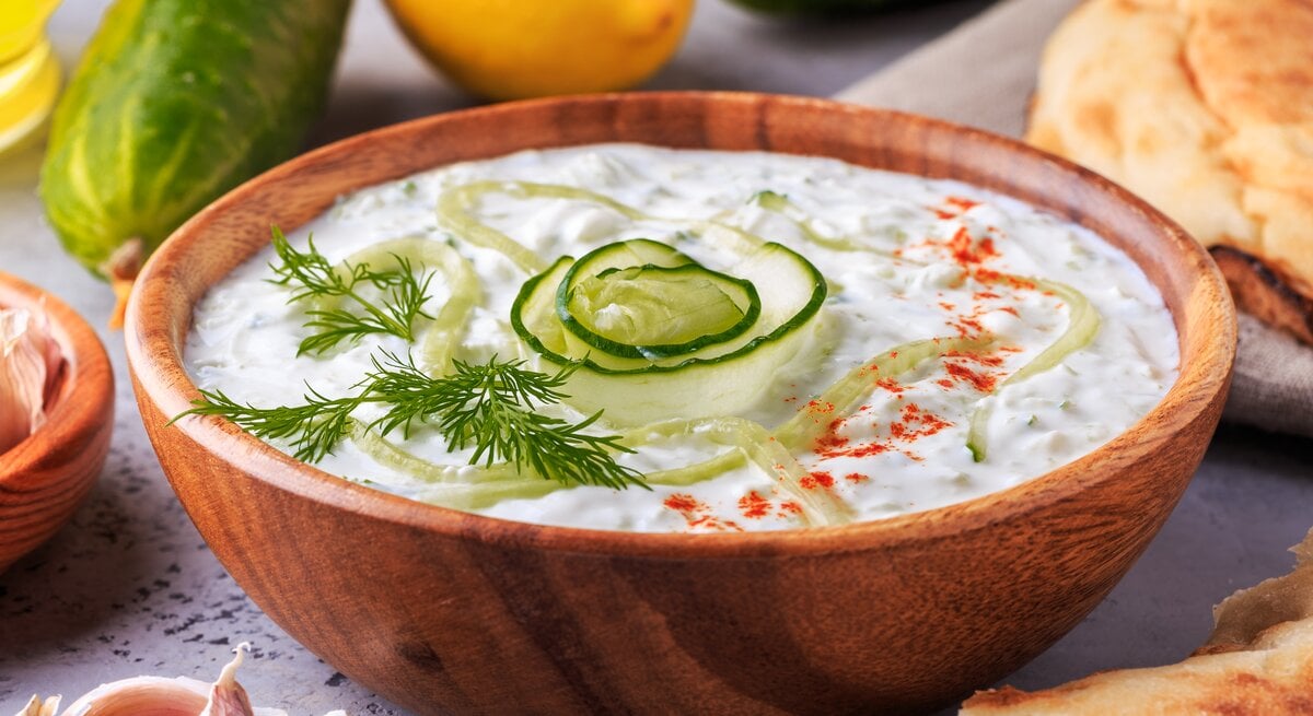 Frischer Zaziki mit Gurke in einer Holzschale. Daneben liegen Gurken, Knoblauch, Zitronen und Brot. 
